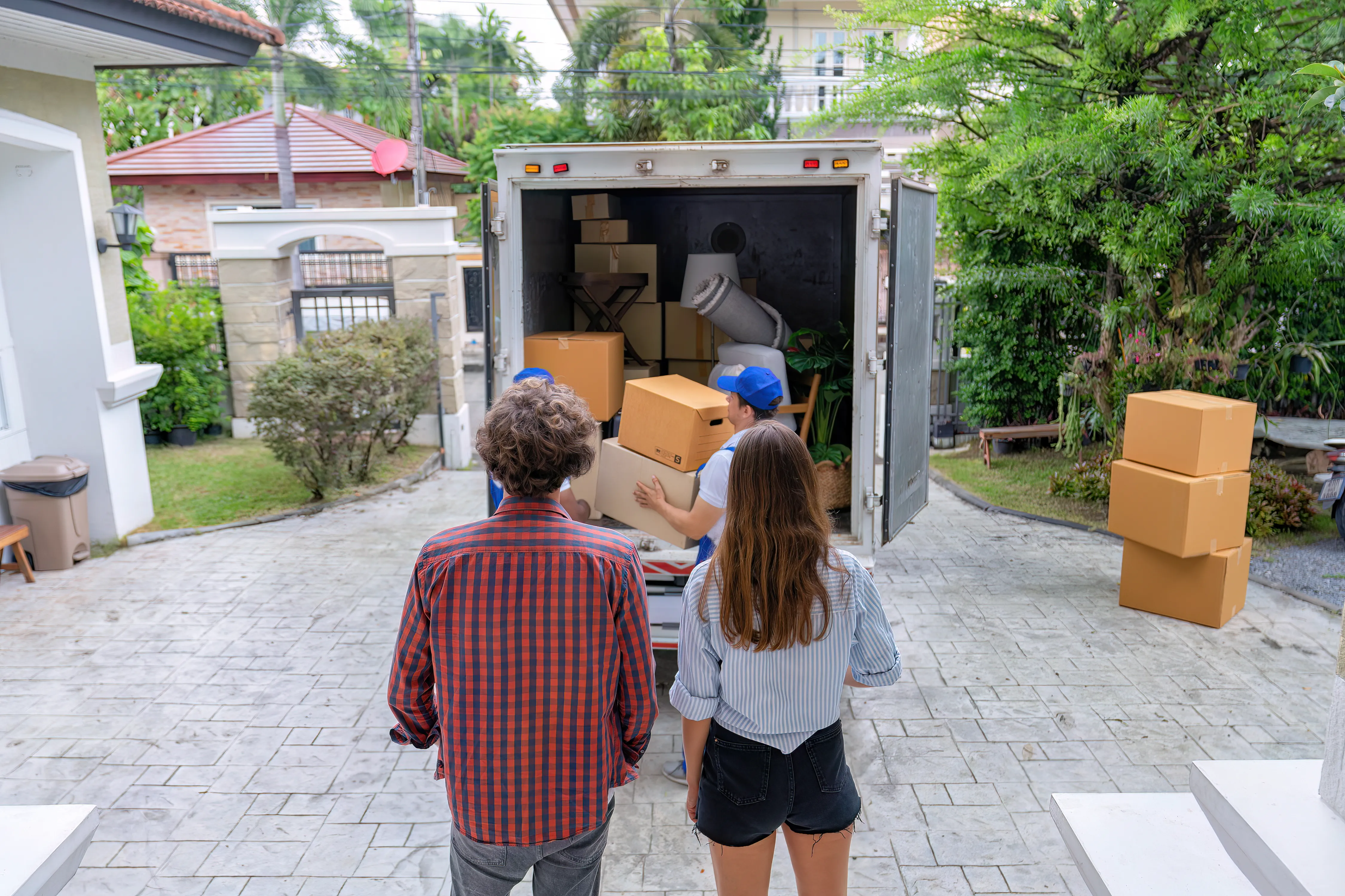 Zwei Personen beobachten Umzugshelfer beim Beladen eines LKWs. Der LKW ist mit Kartons, Teppichen und Möbelstücken beladen.